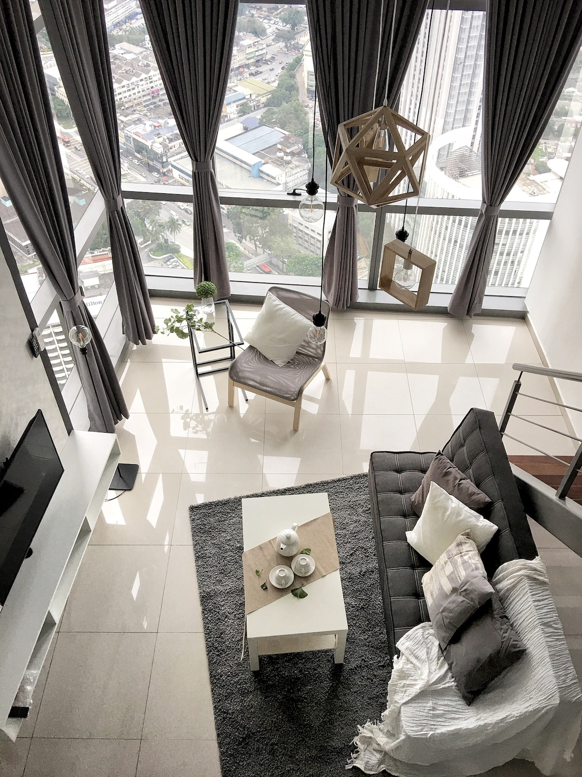 A stylish living area is highlighted by large windows offering a panoramic view of the city. A comfortable gray sofa is complemented by decorative cushions, while a modern coffee table is set with a small plant and two coasters. Natural light brightens the space through subtly draped curtains.