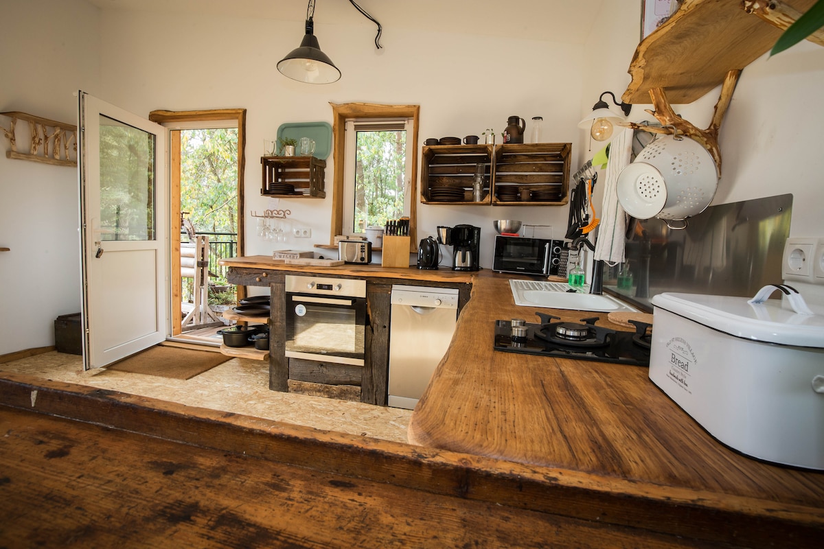A modern kitchen area is displayed with natural wood accents. It features stainless steel appliances, including a microwave and coffee maker, alongside wooden shelving holding kitchenware. Large windows invite natural light, while a door opens to an outdoor space, enhancing the openness of the area.
