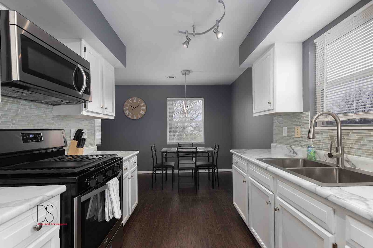 The kitchen features modern appliances, including a stovetop, oven, and refrigerator. White cabinetry complements the marble countertops. A dining table with four black chairs is positioned by a window, allowing natural light to fill the space. A clock adorns the dark gray wall.