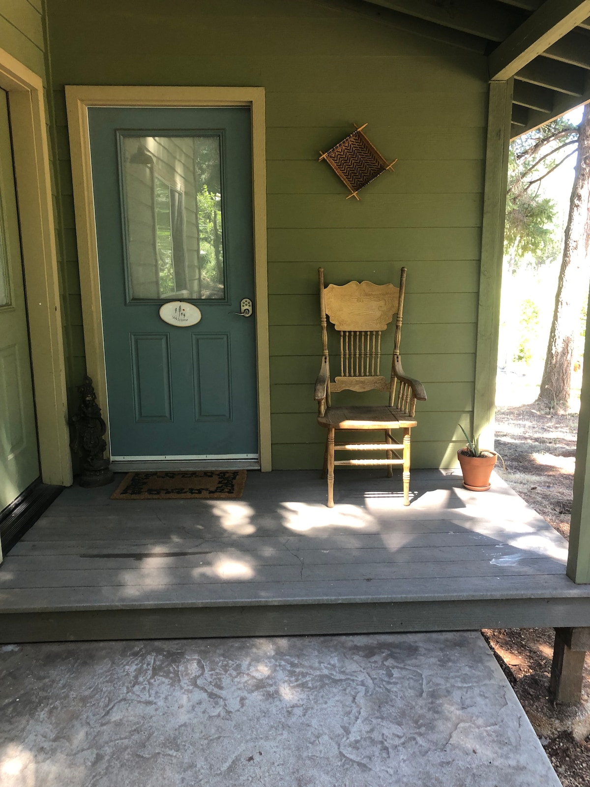 A small front porch features a light green door with a decorative door handle. A wooden chair is positioned adjacent to the door, accompanied by a small potted plant. Natural light filters through the trees, casting shadows on the outdoor flooring.