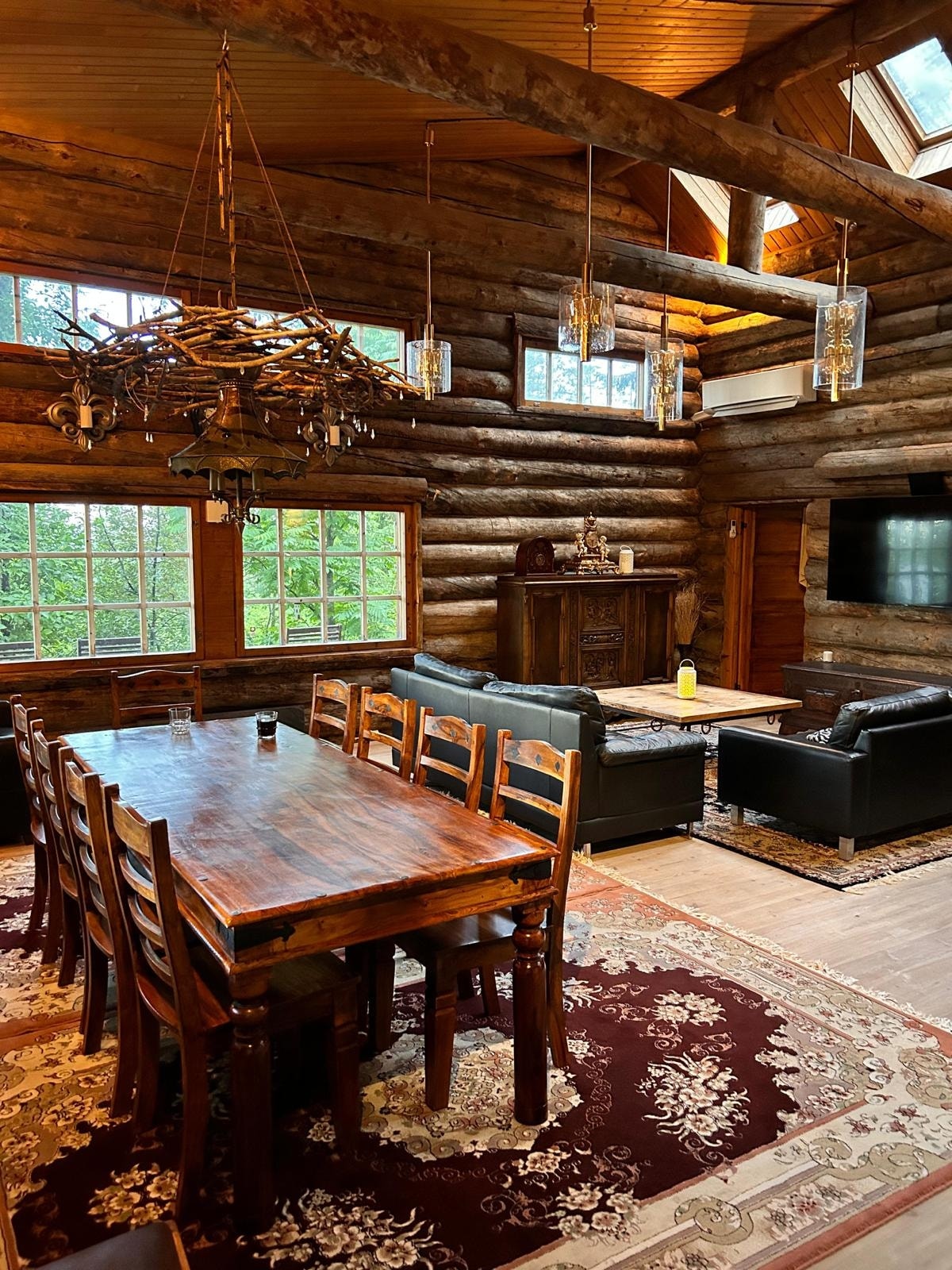 A spacious dining and living area is highlighted by a large wooden dining table surrounded by eight chairs. The room features log walls, large windows allowing natural light, and a decorative chandelier. A comfortable seating arrangement is visible with two black sofas and a wooden coffee table.