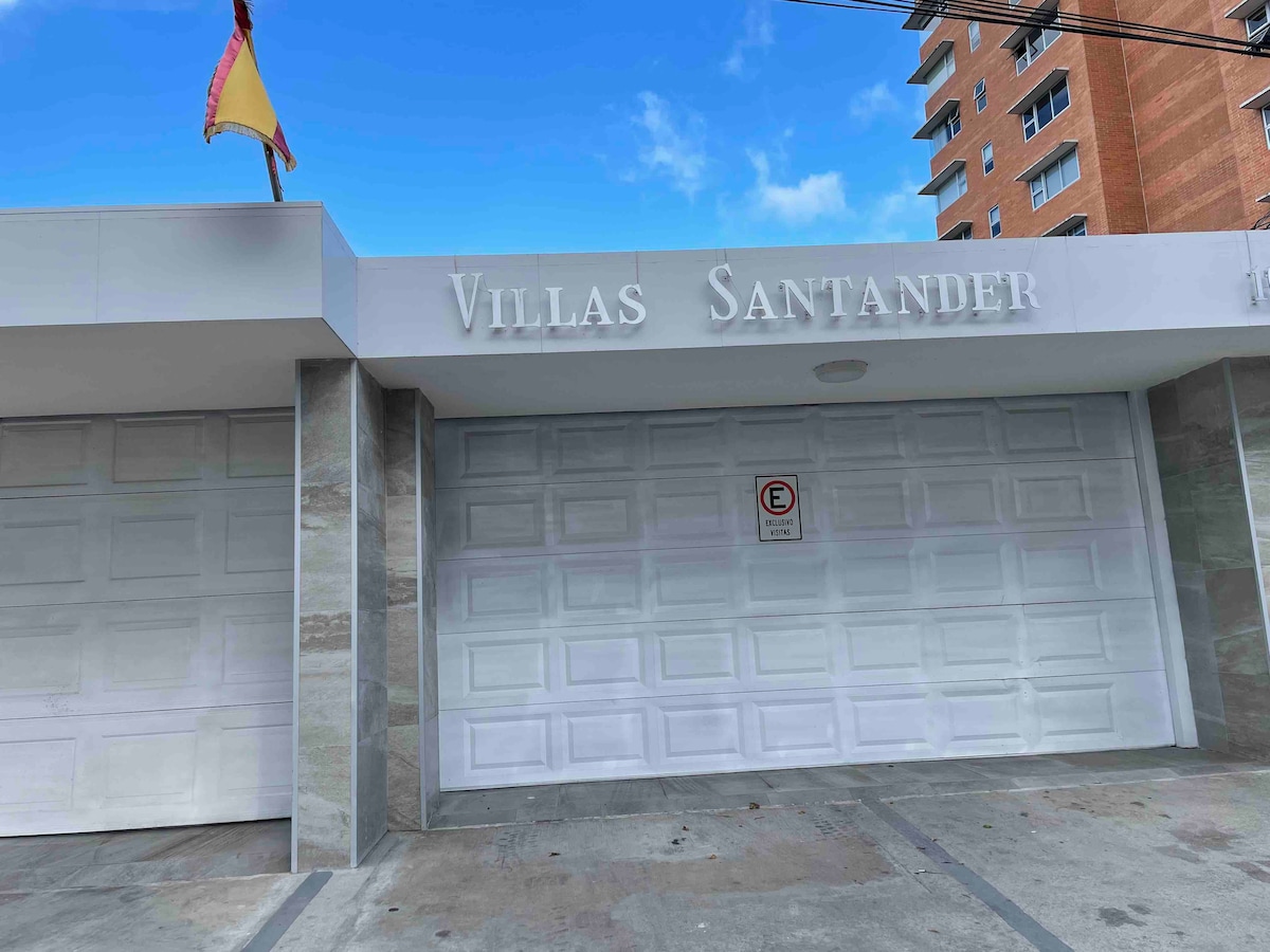 The exterior of Villas Santander is displayed, featuring a modern façade with clear signage. A large, white garage door is visible, complemented by soft grey stone accents. A flag is positioned on the left side under the blue sky.