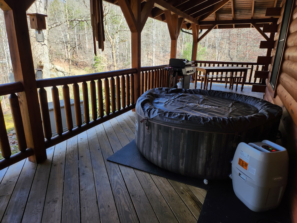 A spacious wooden porch features a hot tub covered for protection and a propane grill nearby. Wooden beams and balusters enhance the rustic charm, with views of the surrounding trees visible in the background.
