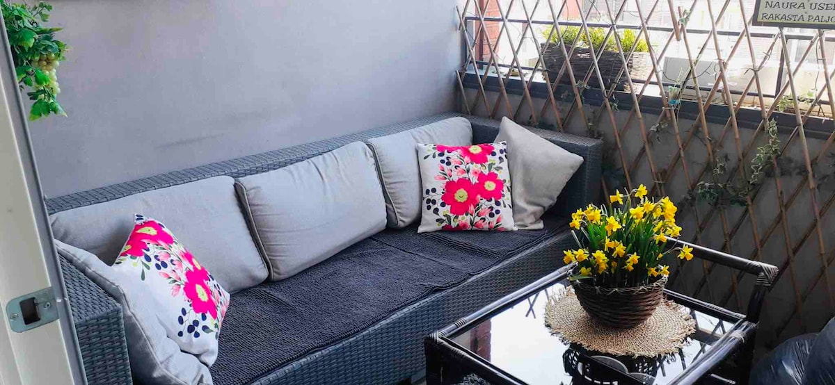 A comfortable seating area is featured on a small patio, showcasing a cushioned sofa adorned with decorative throw pillows. A glass table sits in front, accompanied by a woven basket filled with vibrant flowers, adding a touch of nature to the inviting space.