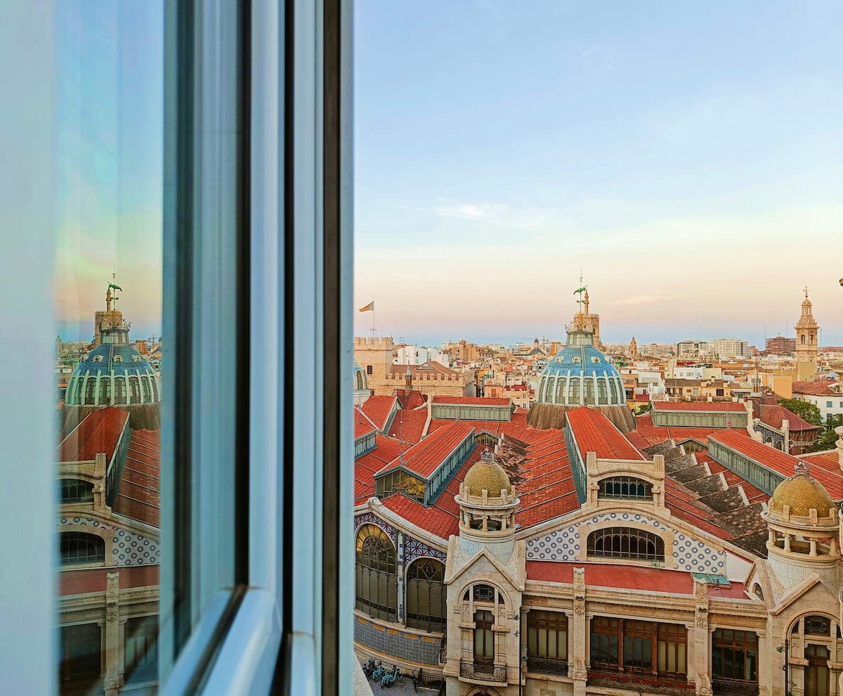 A view of the cityscape is captured through a window, highlighting the iconic rooftops of Valencia. The image features distinctive architectural elements, including the domed structure of the Central Market, under a soft evening sky that transitions in color.