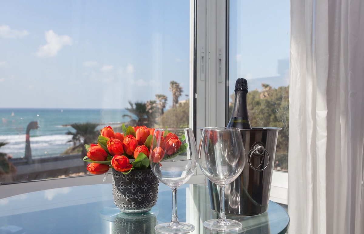 A glass dining table is positioned near a large window, offering a view of the sea. A bouquet of vibrant orange tulips is displayed in a decorative vase, along with a champagne bucket and two glasses, creating an inviting setting.
