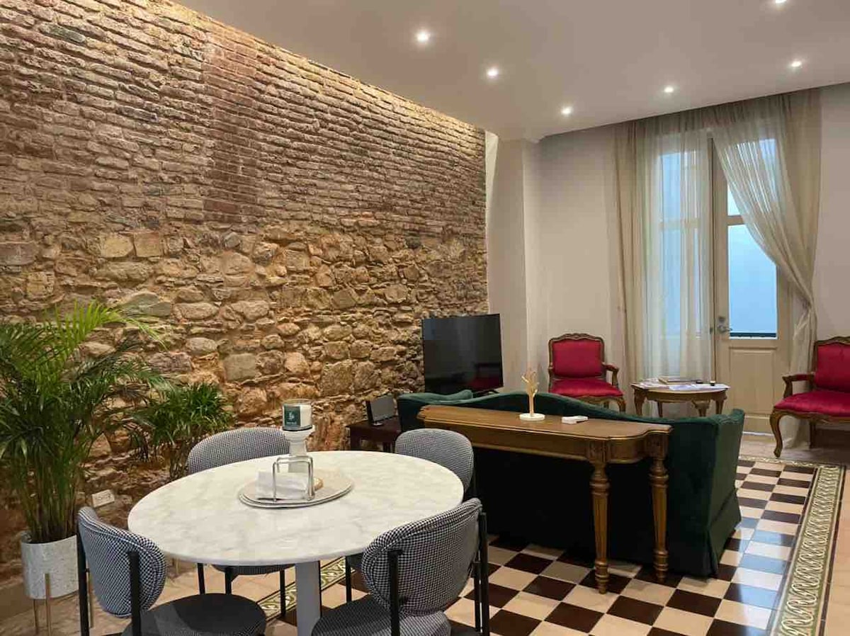 A cozy living area features a round marble dining table surrounded by four upholstered chairs. A rich green sofa is positioned beside an antique table, while a television sits against a backdrop of exposed brick walls. Large windows allow natural light to enter the space.