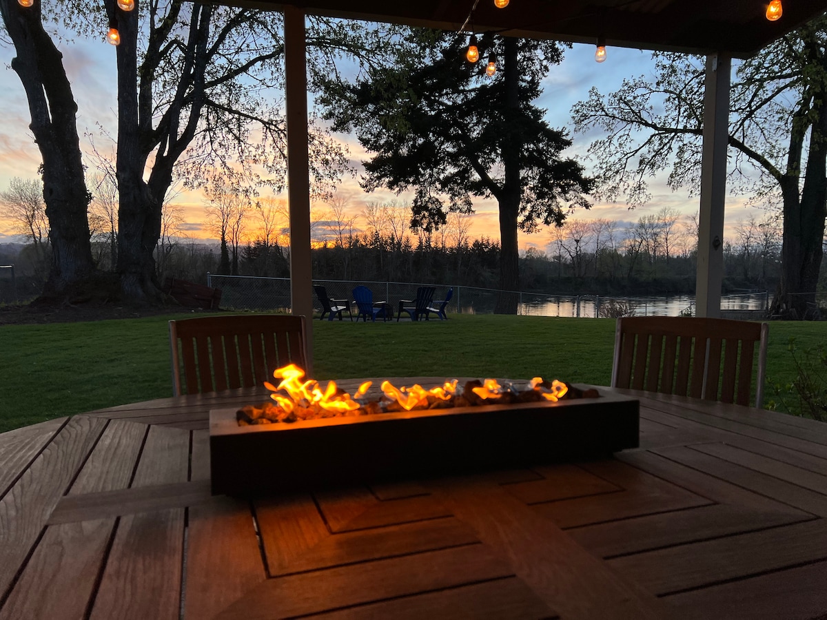 A dining table with a fire feature is positioned outdoors, surrounded by wooden chairs. The backdrop showcases a beautiful sunset over the Willamette River, with vibrant colors reflecting off the water. Trees frame the scene, enhancing the serene atmosphere.