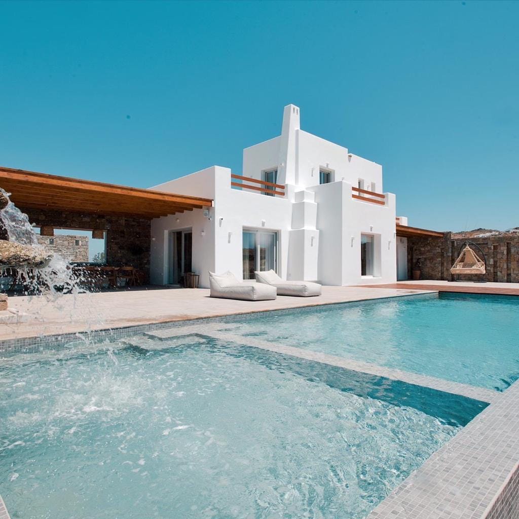 A serene swimming pool is set against a backdrop of a modern villa with a minimalistic design. Light-colored loungers are placed at the edge of the pool, while splashes of water are visible, creating a refreshing atmosphere. Clear skies provide a bright contrast to the structure.