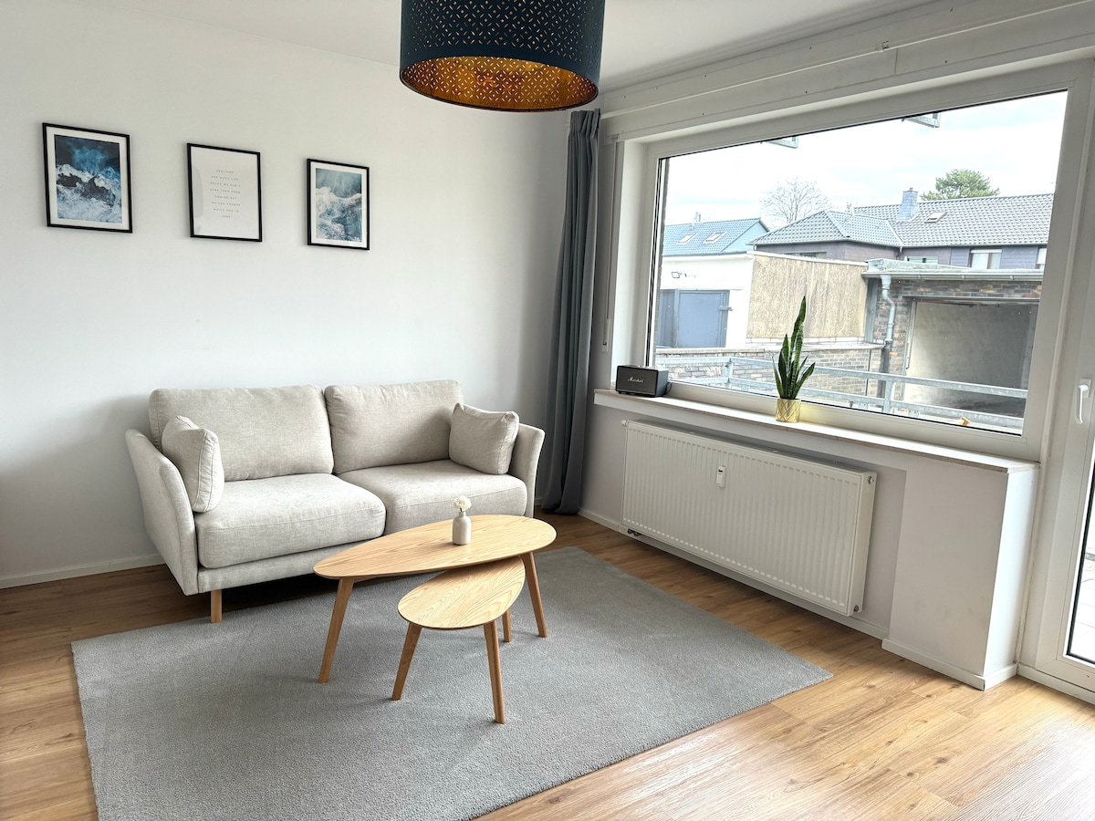 A light-filled living area is furnished with a soft grey couch and a round wooden coffee table. A small potted plant is placed on the window sill, which provides a view of the exterior. Decorative wall art complements the minimalist style.