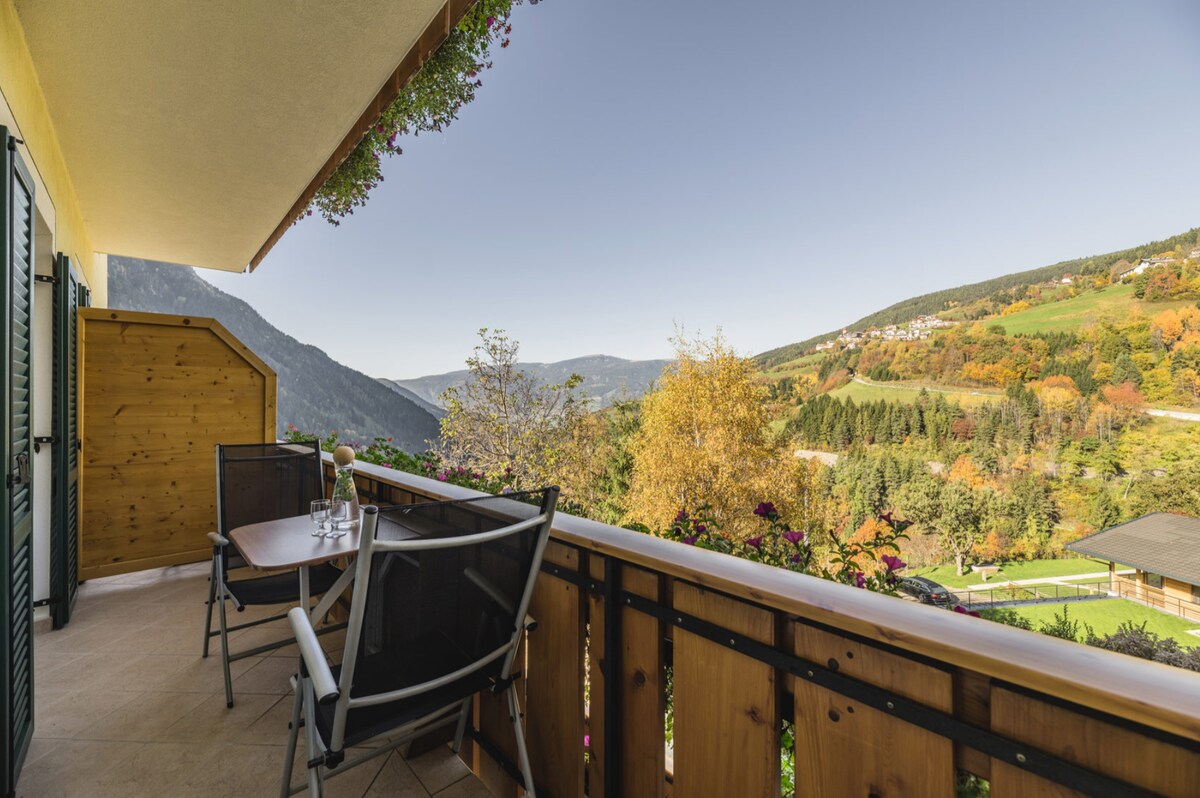 A balcony is visible, featuring a wooden railing with potted plants along the edge. A small table and two chairs are positioned to enjoy the expansive views of the mountains and valley, where autumn foliage creates a colorful landscape against a clear blue sky.