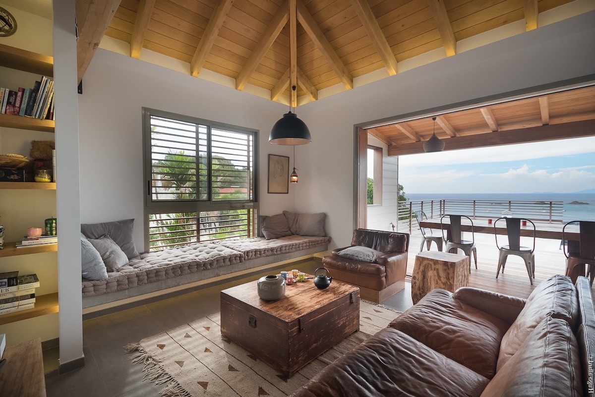 A spacious living area features a high wooden ceiling and large windows that offer ocean views. A comfortable seating arrangement includes two leather sofas and a wooden coffee table. A cozy window nook provides additional seating, enhancing the inviting atmosphere of the space.