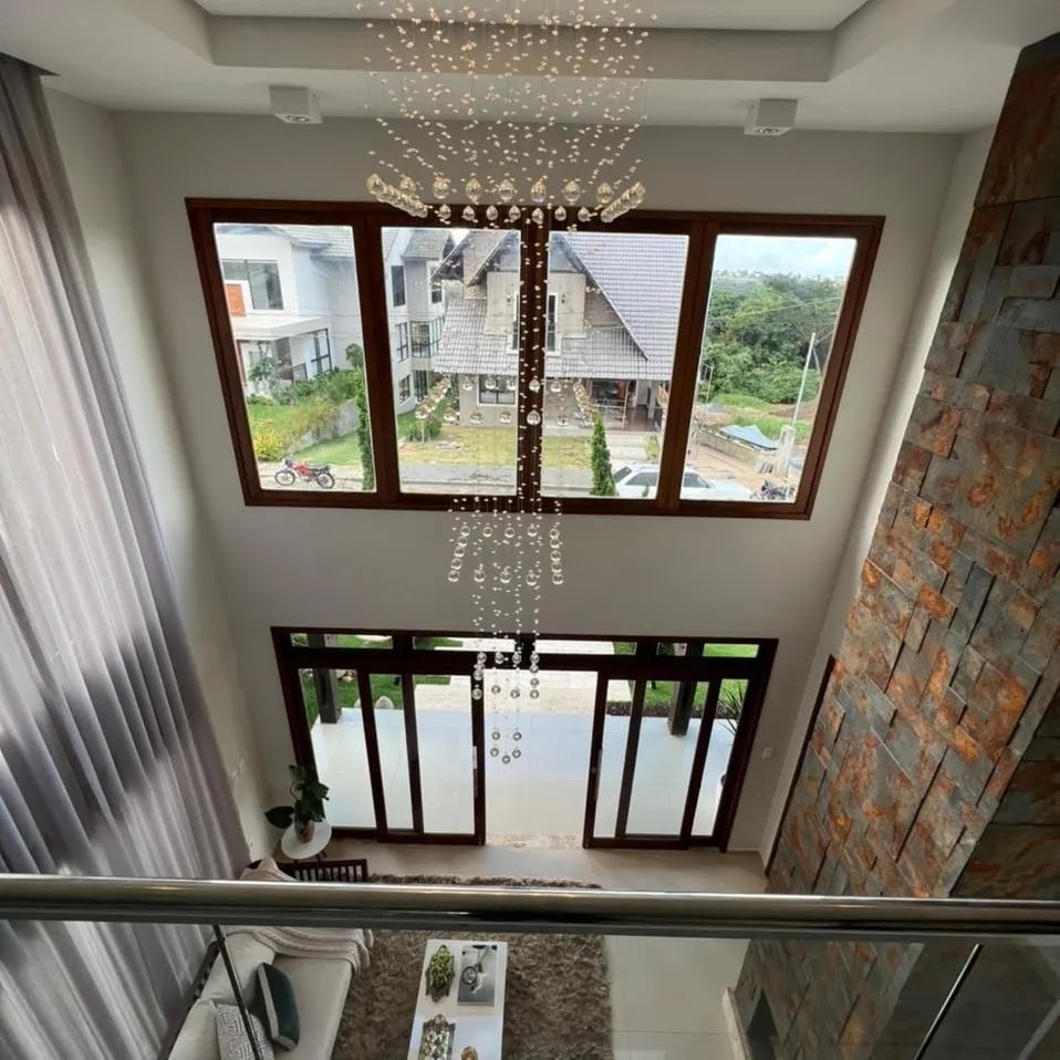 A spacious interior is highlighted by large windows, allowing natural light to flood the area. A modern chandelier hangs gracefully from the ceiling, while a soft rug complements the contemporary furnishings below, creating a welcoming ambiance. Lush greenery can be seen outside.