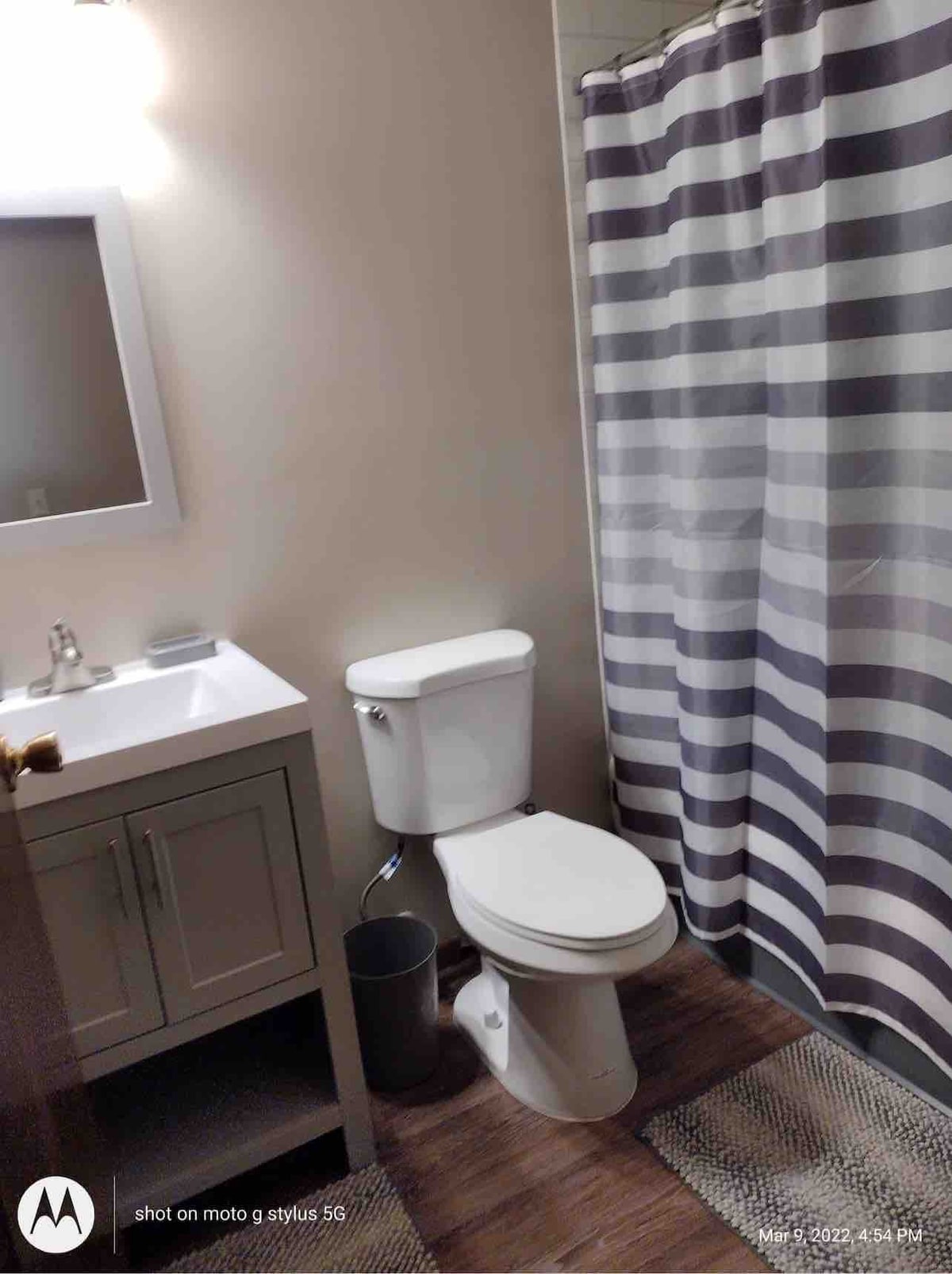 A clean bathroom features a modern vanity with a rectangular mirror above. A neutral-toned shower curtain with gray stripes and a woven rug complement the space. A white toilet is positioned alongside a round wastebasket, enhancing the overall functionality of the area.