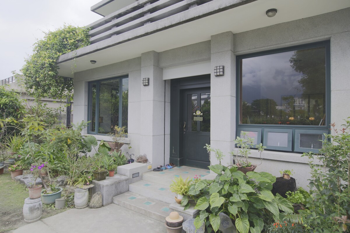 The entrance features a modern facade with large windows and double doors framed by lush greenery. Potted plants line the pathway, showcasing a variety of flora. The outdoor space is well-maintained, creating an inviting access point to the home.