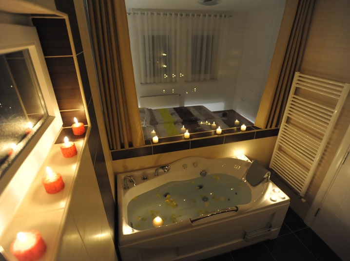 A bathroom features a spacious whirlpool bathtub surrounded by softly flickering candles. A large mirror reflects a cozy sleeping area with soft bedding and curtains filtering ambient light. Modern fixtures and a towel warmer add to the contemporary design.