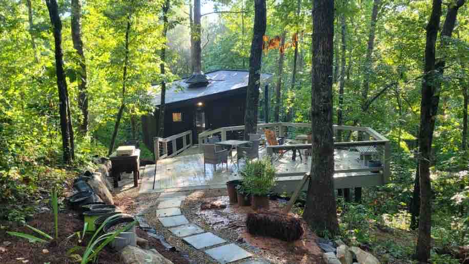 A winding stone pathway leads to a modern treehouse nestled among lush greenery. A spacious deck surrounds the building, featuring a seating area and grill. Towering trees provide a natural canopy, enhancing the secluded, tranquil atmosphere of the setting.