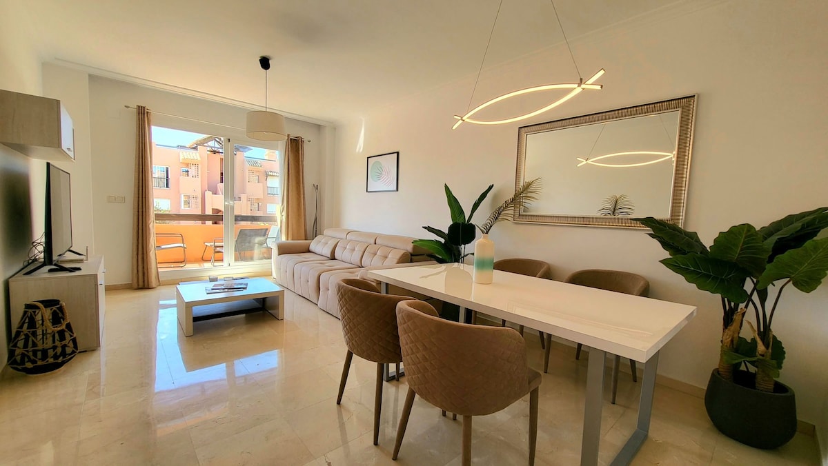A modern living space features a light-colored sectional sofa, a sleek coffee table, and a dining table with stylish chairs. Large windows allow natural light to fill the room, and decorative plants add a touch of greenery. A minimalist chandelier provides soft illumination.