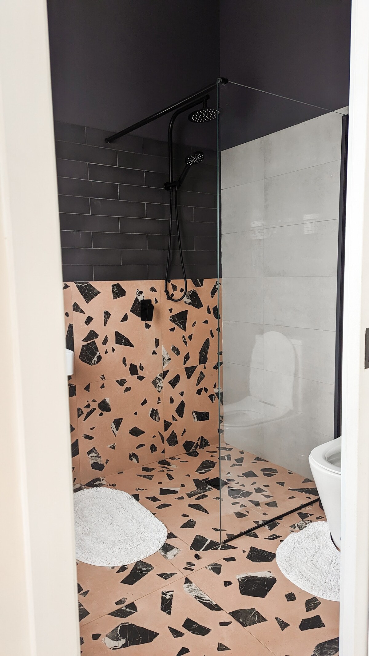 A modern bathroom is presented with a glass shower enclosure featuring a sleek rainfall showerhead. The floor displays a unique pattern with light and dark tiles, while soft circular bath mats are positioned in front of the shower and toilet.