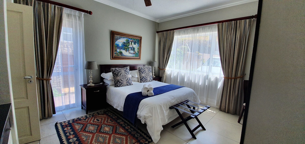 A well-lit room features a spacious bed adorned with decorative pillows. Two large windows allow natural light to fill the space, which is complemented by neutral-toned walls and elegant drapery. A small side table with lamps and artwork can be seen, enhancing the overall decor.