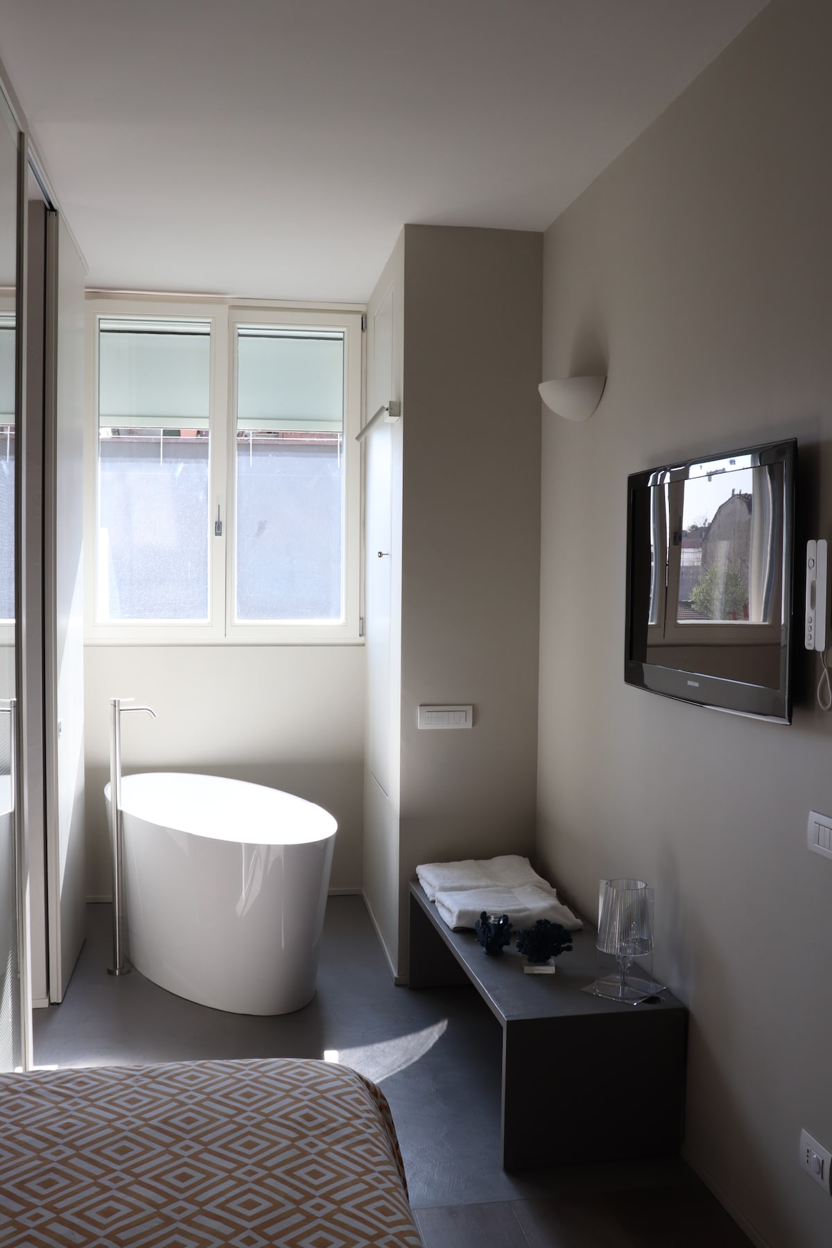 A modern bathroom is depicted with a freestanding tub positioned near a window, allowing natural light to illuminate the space. A wall-mounted television is visible alongside a sleek cabinet featuring folded towels and decorative items, all set against neutral-toned walls.