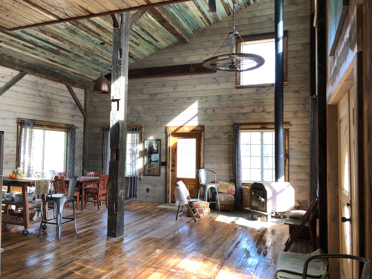 A spacious interior features high, rustic wooden ceilings and natural light filtering through large windows. Exposed beams and salvaged materials enhance the character of the room. Various seating options are visible, along with a wood stove and a dining area that encourages gathering.
