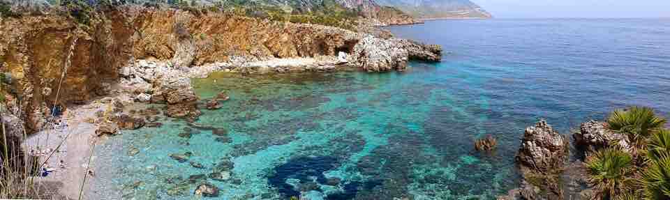 A tranquil coastal scene is presented, featuring clear turquoise waters gently lapping against rocky shores. Lush greenery lines the cliffs, creating a natural backdrop. The shoreline is dotted with smooth stones and pebbles, inviting exploration of the peaceful beach area.