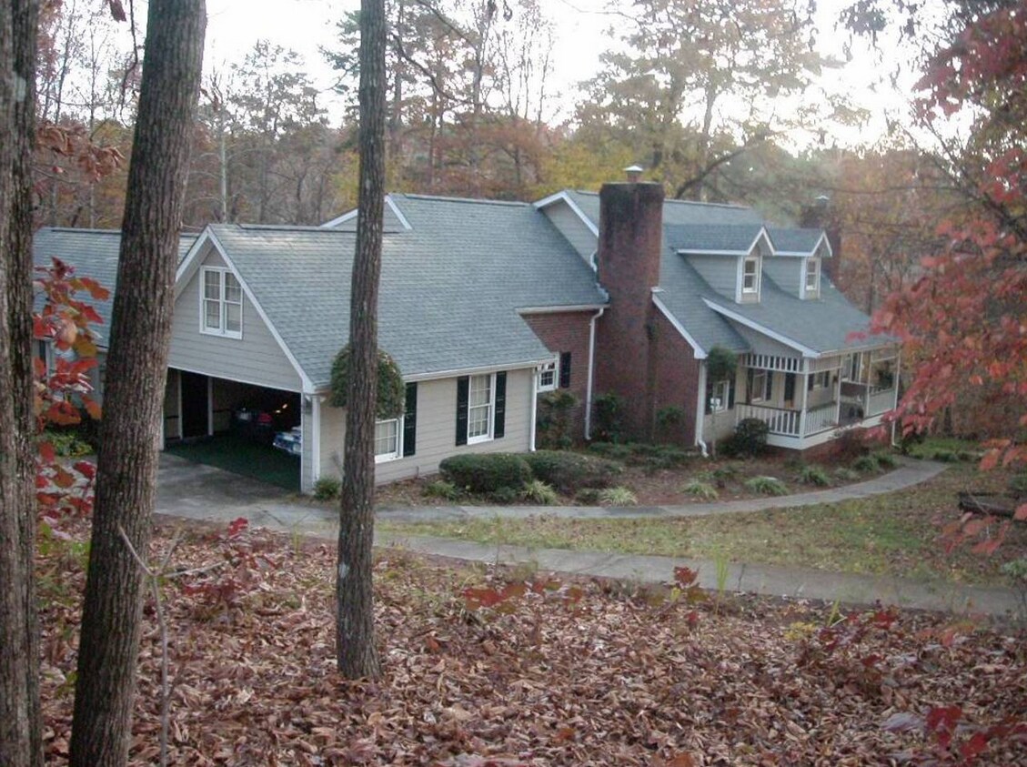 The exterior of a charming two-story home is surrounded by trees, featuring a sloped roof and a brick chimney. A porch is visible along the side, while a carport is integrated into the structure. Autumn leaves blanket the ground, enhancing the wooded setting.