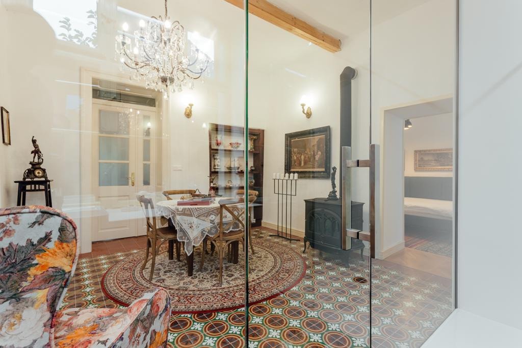 A dining area is shown with a wooden table surrounded by four chairs, illuminated by a chandelier overhead. A decorative cabinet and a wood stove add character, while patterned tiles create a vibrant flooring backdrop. A doorway leads to another room in the background.