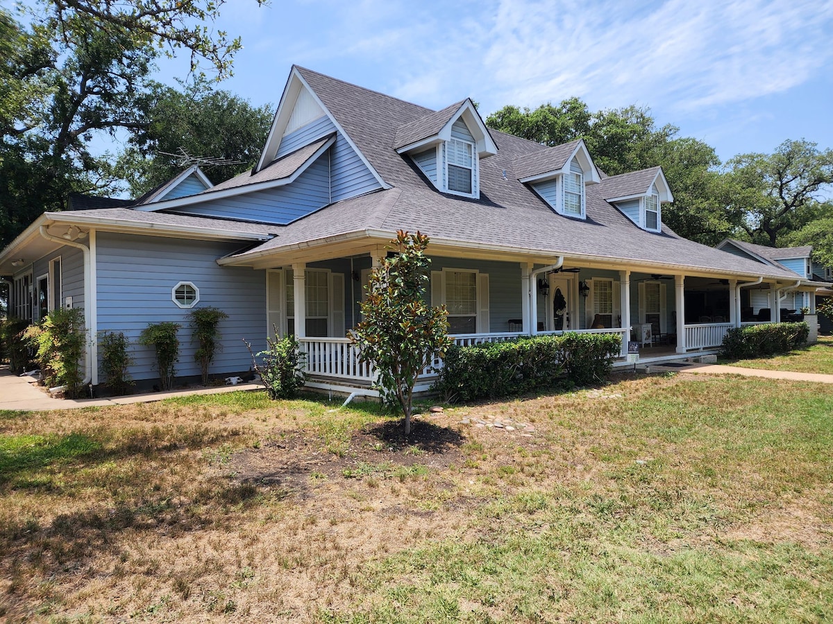 A charming two-story home is surrounded by lush greenery and well-maintained landscaping. A spacious porch lined with white railings invites relaxation, and multiple gabled roofs enhance the architectural interest. The blue exterior harmonizes with the natural surroundings, complementing the serene, open spaces of the property.