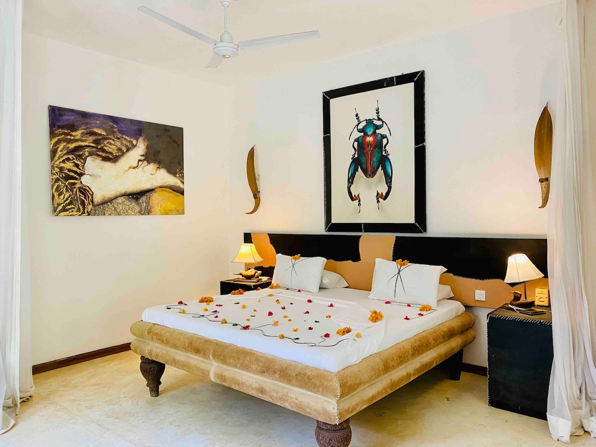 The sleeping area features a spacious bed adorned with flower petals, complemented by unique artwork on the walls. A ceiling fan is positioned above, and bedside tables with lamps provide soft lighting. Textured elements add an interesting aesthetic to the neutral-toned decor.