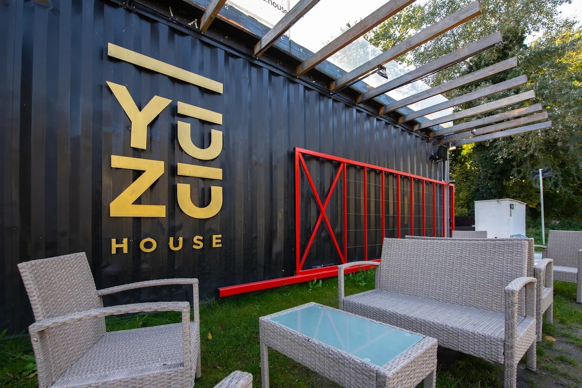The exterior of YUZU House is featured, constructed from a converted shipping container. The logo is prominently displayed in gold lettering against a black background. A shaded seating area is visible, furnished with light wicker chairs and a glass-topped table, surrounded by green foliage.