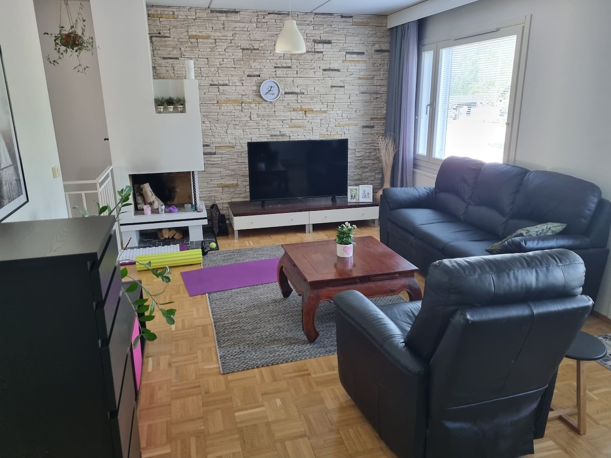 A cozy living area features a black leather sofa and armchair, arranged around a wooden coffee table. A television is mounted on a light-colored stone wall, complemented by a decorative clock. Natural light enters through the window, enhancing the warmth of the space.