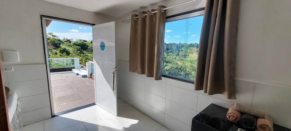 A well-lit room is displayed, featuring a large window that offers views of lush greenery. A door leads to an outdoor area, while a compact space includes a countertop and two rolled towels. Light-colored curtains frame the window, contributing to a bright ambiance.