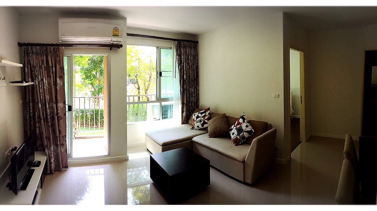A bright living area features a comfortable sectional sofa adorned with decorative cushions. Large windows offer natural light and views of greenery outside. A flat-screen TV is mounted on the wall, and a dark coffee table occupies the center of the space, complemented by modern decor.
