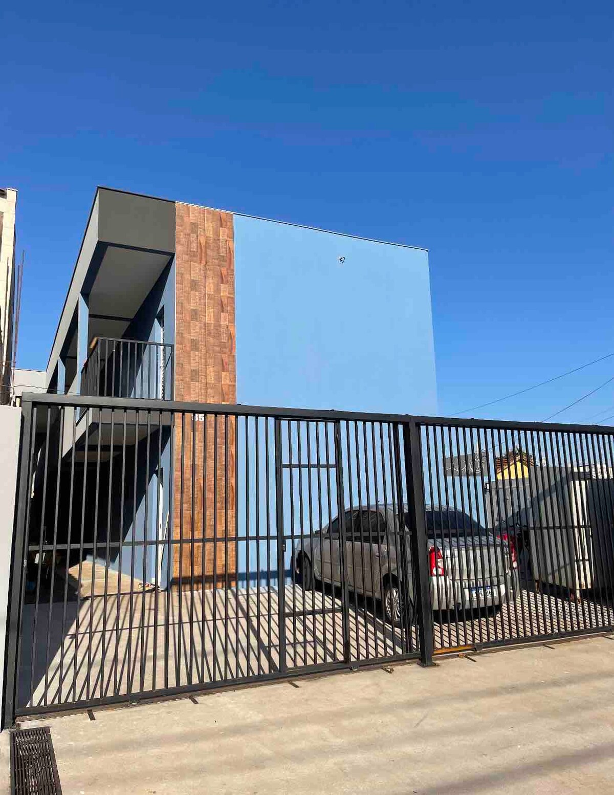 A modern two-story building is presented behind a black metal gate. The exterior features a combination of blue and textured brick accents. A parked vehicle is visible in the spacious driveway, under a clear blue sky.