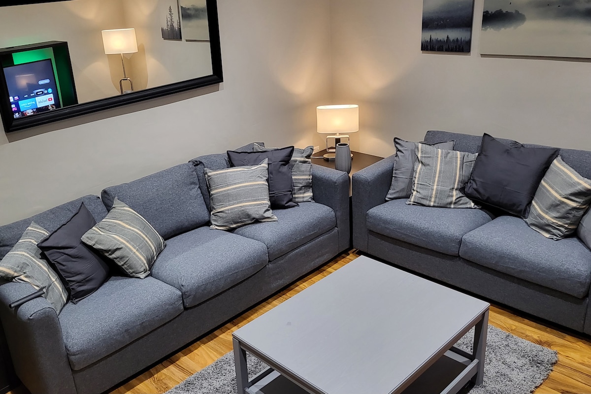 A comfortable seating area features two grey sofas adorned with various patterned cushions. A light-colored coffee table sits on a soft rug, while a flat-screen TV and a lamp provide soft illumination against the neutral wall backdrop.