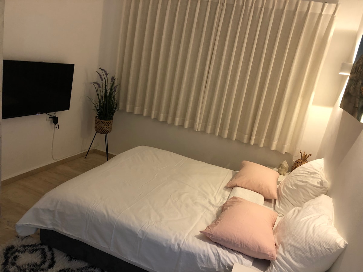 A spacious sleeping area features a large, neatly made bed with light bedding and decorative pillows. A wall-mounted television is positioned opposite the bed, and soft, sheer curtains allow natural light to filter in, creating a calm ambiance.