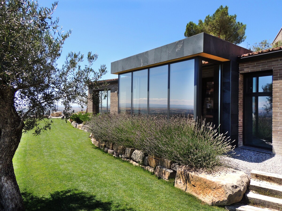 A modern structure features large glass windows that reflect the surrounding landscape. The exterior is surrounded by a well-maintained lawn and decorative stones, with olive trees and lavender enhancing the natural beauty of the area.