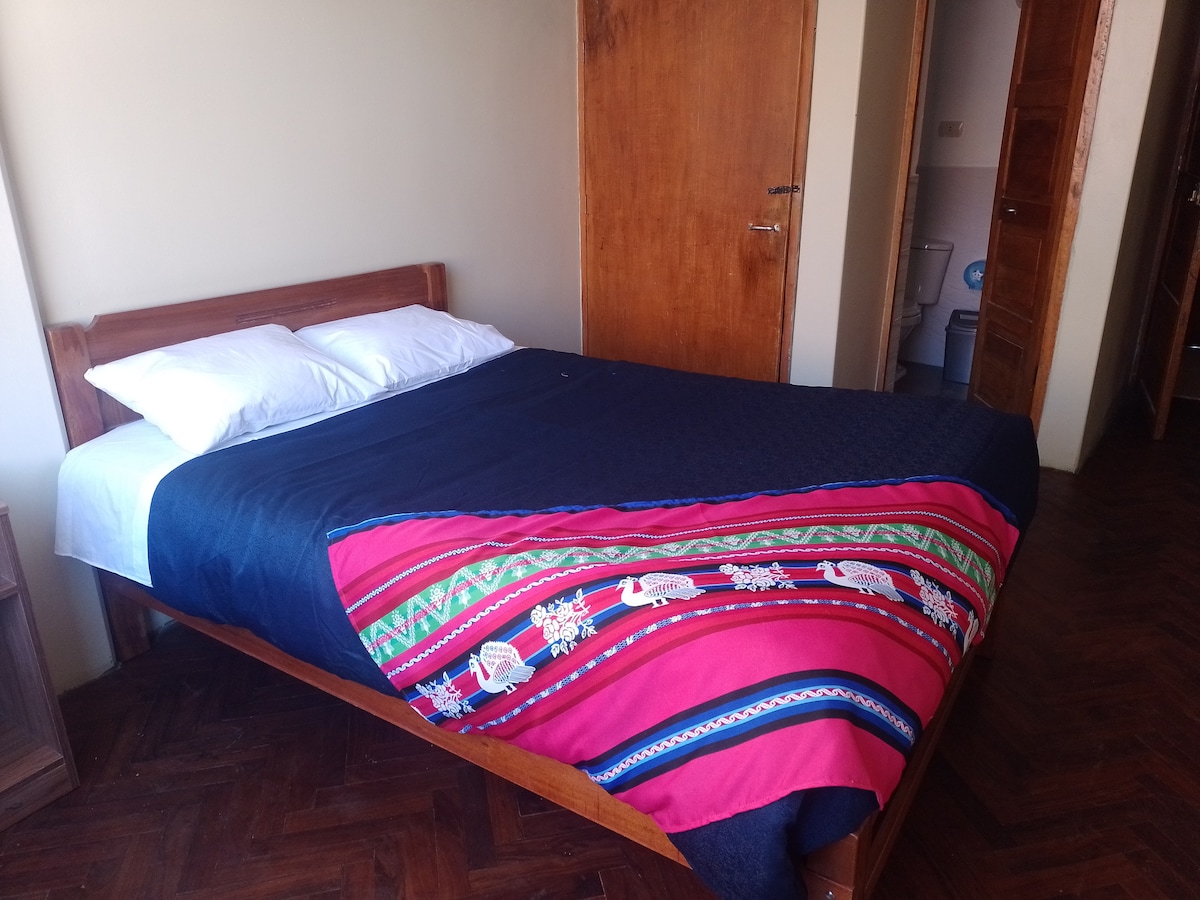 A spacious bed is dressed in a colorful blanket featuring traditional patterns. Natural light enters through a nearby window, illuminating the room. Wooden furniture, including bedside tables, complements the warm tones of the walls and floor.