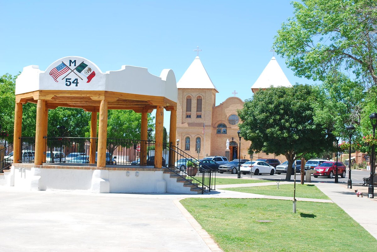 A public gazebo is positioned prominently in the foreground, featuring wooden beams and decorative sign displaying US and Mexican flags. In the background, a historic church with twin steeples stands under a clear blue sky, surrounded by lush green trees and parked cars along a pathway.