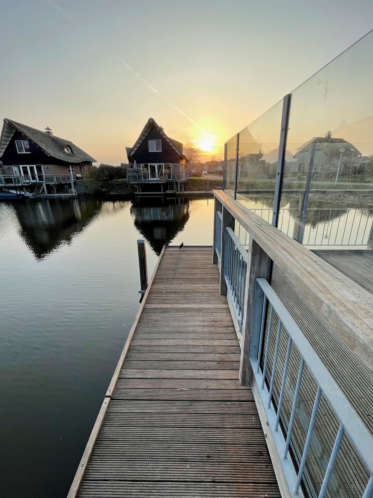 A wooden walkway extends over calm water, leading towards two sloped-roof houses. The warm glow of the setting sun reflects on the water's surface, creating a serene atmosphere. Glass railings provide a clear view of the surroundings, enhancing the peacefulness of the scene.