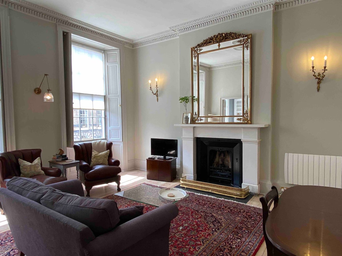 The living area features tall windows allowing ample natural light. A large mirror is mounted above a decorative fireplace, with two armchairs and a comfortable sofa arranged around a small coffee table. Soft lighting is provided by wall sconces.