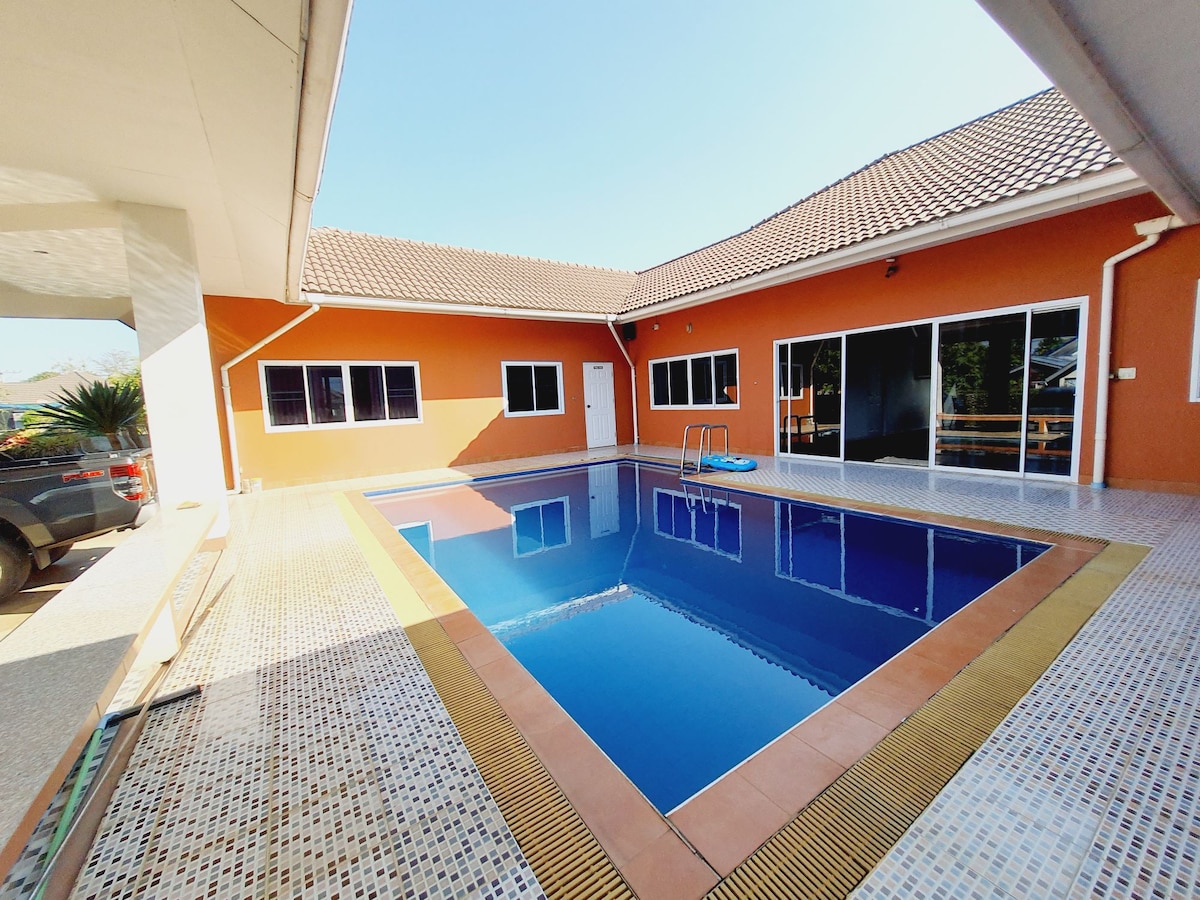 A rectangular pool is surrounded by a spacious patio. The vibrant orange exterior of the building contrasts with the clear blue water. Large glass doors allow views of the pool area, and several windows provide natural light. A few lounge chairs are positioned near the water.