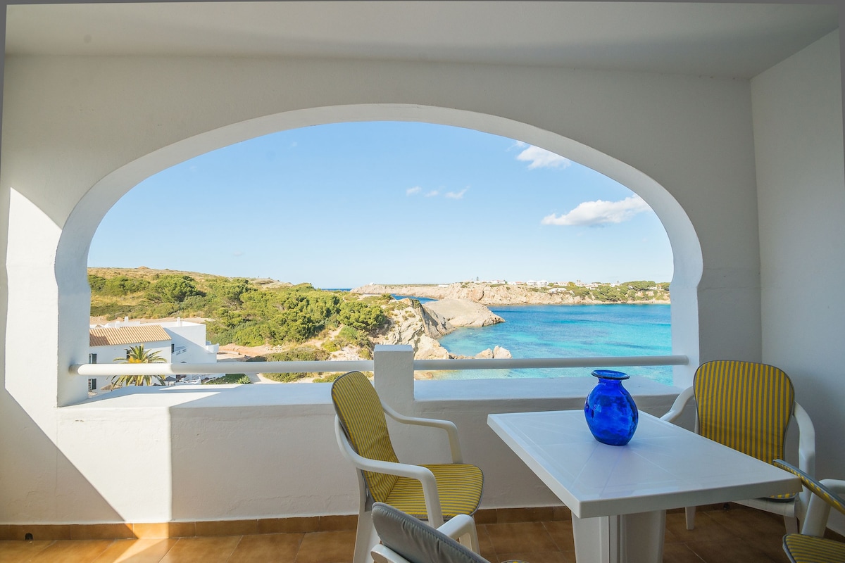 A covered balcony features a table and two chairs, with a vibrant blue vase on the table. An expansive view of the turquoise sea and rocky coastline is visible through the archway, framed by white walls and airy light.