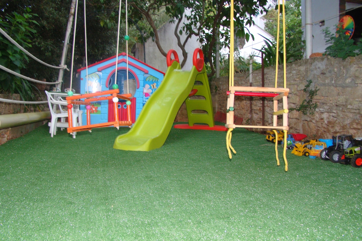 A private playground is featured in the outdoor space, with a bright green slide and swings. Colorful play structures are set against a lush backdrop, while a small wooden playhouse adds an element of fun. Nearby, toy vehicles are arranged on the ground.