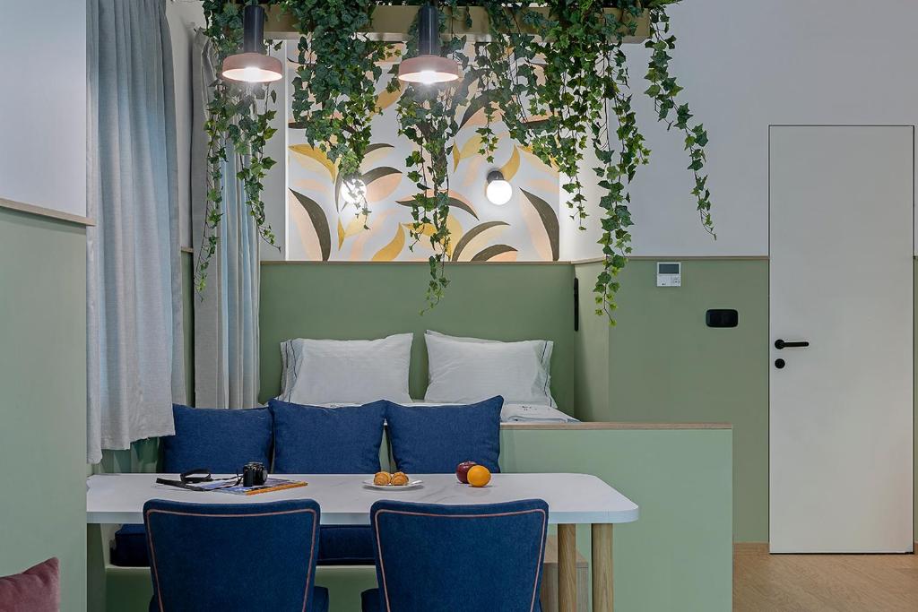 A cozy seating area features a white table with dark blue chairs, complemented by wall art and greenery suspended above. The sleeping area, partially visible in the background, includes a comfortable double bed with white bedding against a light green wall.