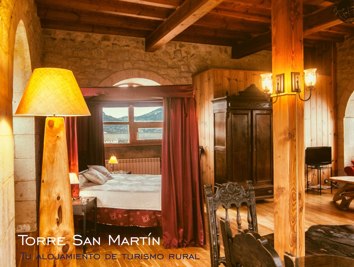 A cozy suite features a spacious bed draped with rich red curtains. A wooden beam ceiling and rustic furnishings, including an antique wardrobe, create a warm environment. A small seating area and a television are positioned to offer comfort and relaxation, with scenic valley views visible through the windows.