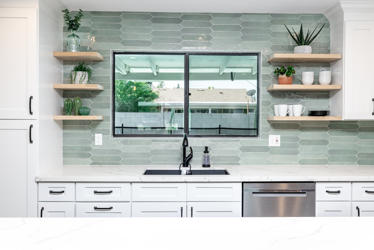 The kitchen features a sleek design with white cabinetry and stainless steel appliances. Open shelving displays decorative glassware and potted plants against a modern green tile backsplash. A large window provides natural light and views of the outdoor area.