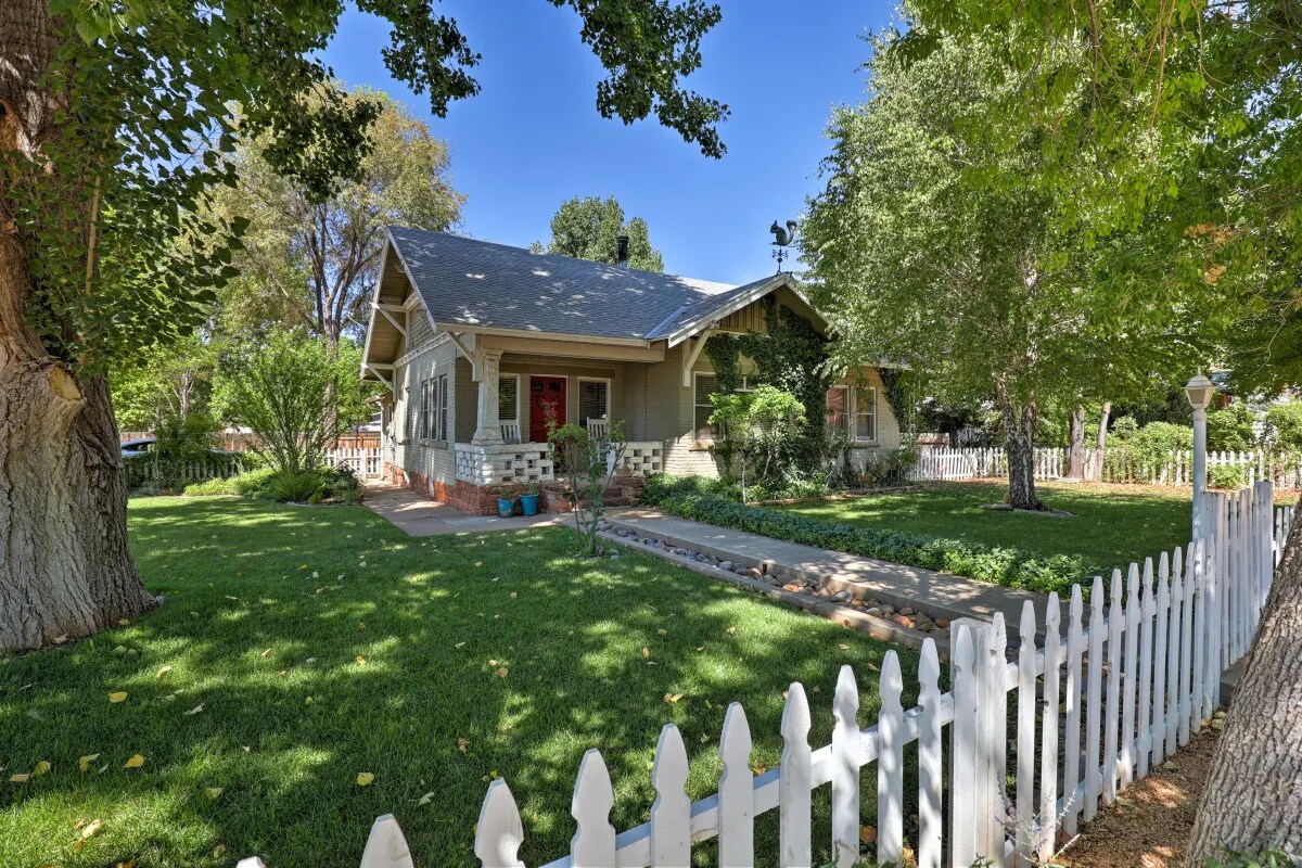 A charming cottage is framed by a white picket fence and lush greenery. Large trees provide shade in the front yard, and a well-maintained pathway leads to the entrance, complemented by colorful flower beds on either side.